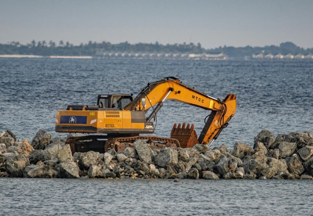 An excavator works on the rocky shoreline, part of a coastal construction project by the sea.