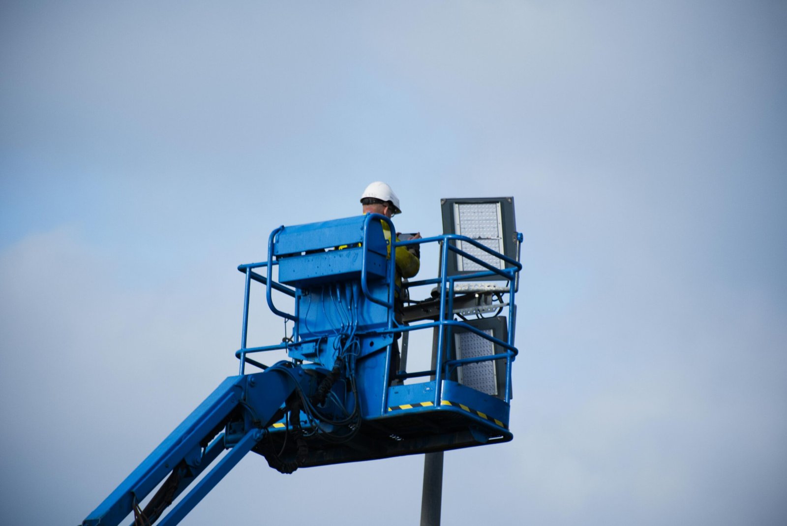 An industrial worker wearing a hard hat operating a lift platform for maintenance tasks in an outdoor setting.
